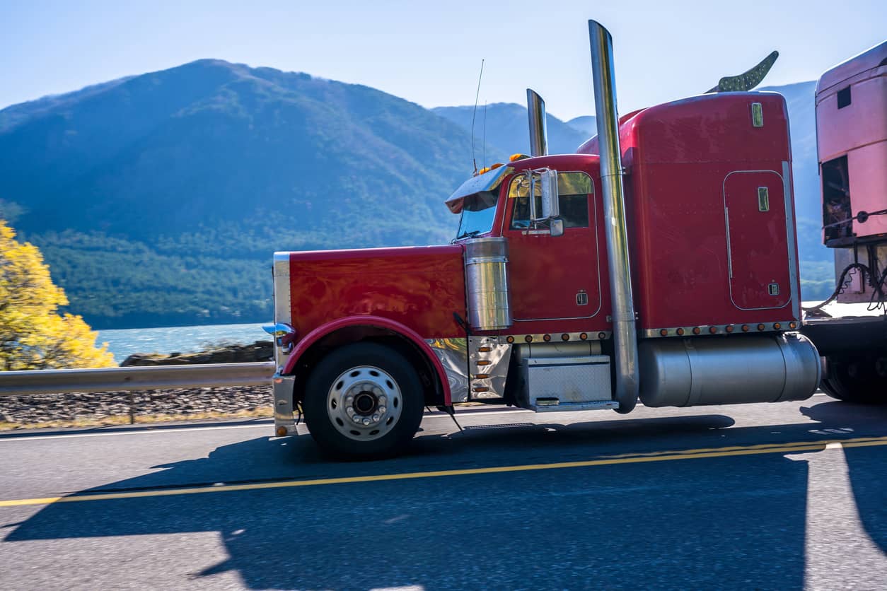 Red classic big rig semi truck with chrome parts driving on the road along the river